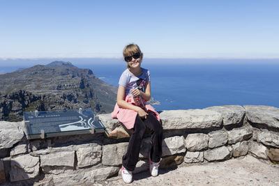 Full length of girl sitting on rock against sea and clear sky