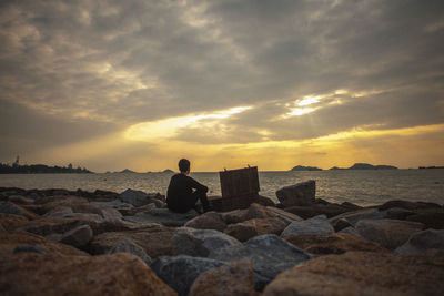 Man with box sitting at rocky sea shore against cloudy sky during sunset