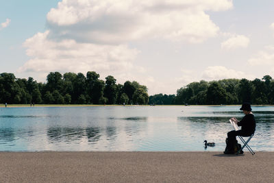 Side view of person holding paper while sitting by bird in lake against sky