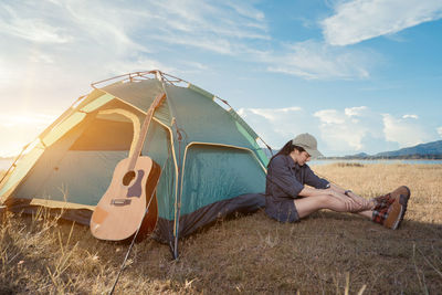 Side view of young woman sitting on land by tent against sky