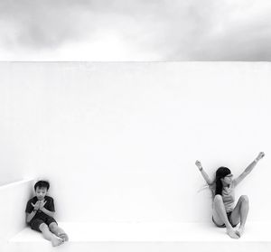 Full length of siblings sitting by white wall against cloudy sky