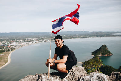 Portrait of smiling young man holding flag while crouching on mountain against sea at beach