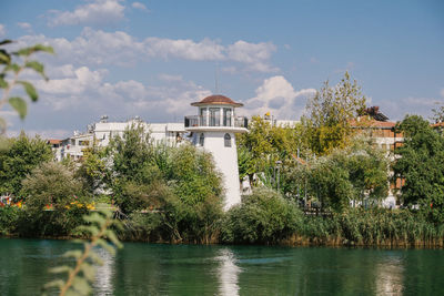 Trees and buildings by river against sky