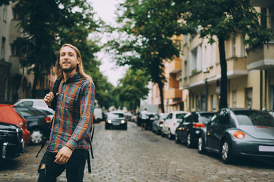 Young man carrying backpack while walking on cobbled street amidst cars in city