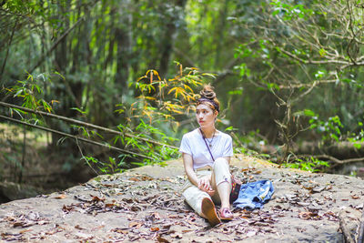 Thoughtful young woman sitting on rock against trees in forest