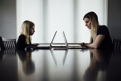 Side view of mother and daughter using laptops on table while sitting at home