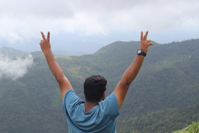 Rear view of man with arms raised gesturing peace sign against mountains