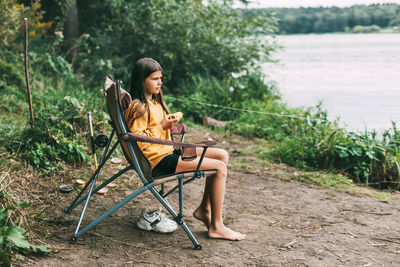 A teenage girl in a bright yellow sweater is sitting in a camping chair on the shore of the lake 