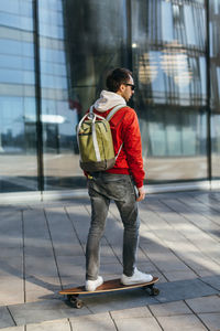 Stylish hipster in red jacket, sunglasses and with backpack riding on longboard. selective focus.