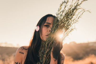 Photo of a beautiful dark-haired woman with tattoos intimately hugging herbs to face at golden hour.