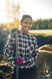 Portrait of smiling mid adult female farmer holding shovel with pigs in background at organic farm