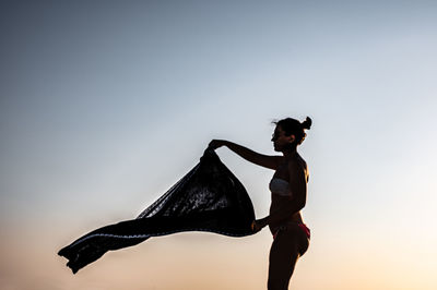 Side view of woman holding scarf against clear sky at sunset