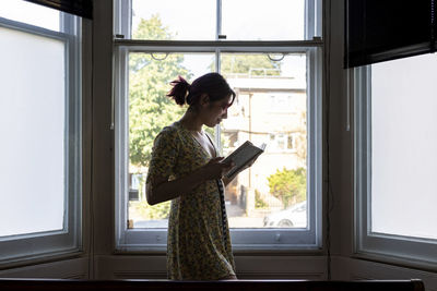 Smiling woman reading book standing in front of window