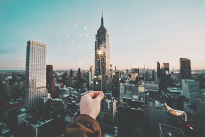 Cropped hand of person holding lit sparkler against cityscape