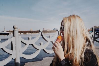 Side view of smiling young woman eating ice cream on pier by sea against sky