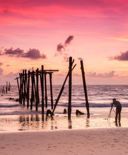 Photographer photographing abandoned pier on beach against sky during sunset