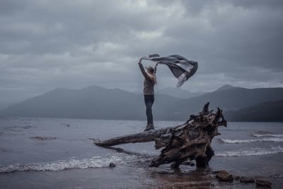 Man standing on driftwood at beach against sky