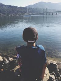 Rear view of boy sitting on rocks at lakeshore