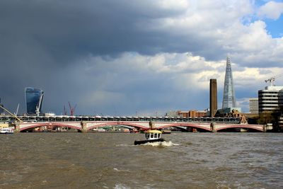 Boat moving on river by bridge against cloudy sky