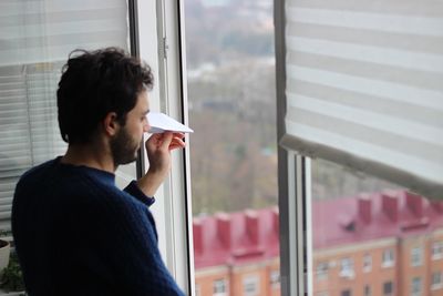High angle view of man holding paper airplane while standing by window at home