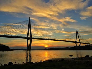 Silhouette of suspension bridge over river against cloudy sky