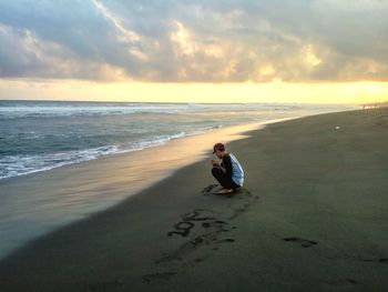 Man sitting on shore at beach against sky during sunset