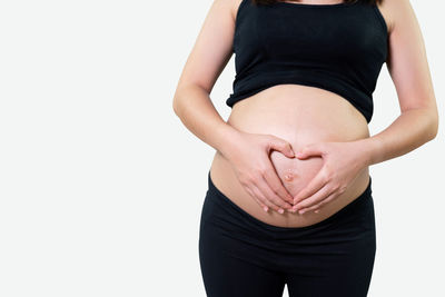 Midsection of pregnant woman making heart shape on stomach with hands over white background