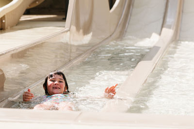 From above little girl with wet dark hair riding on water slide while spending sunny summer day in aqua park