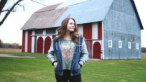 Young woman looking away while standing on field against barn