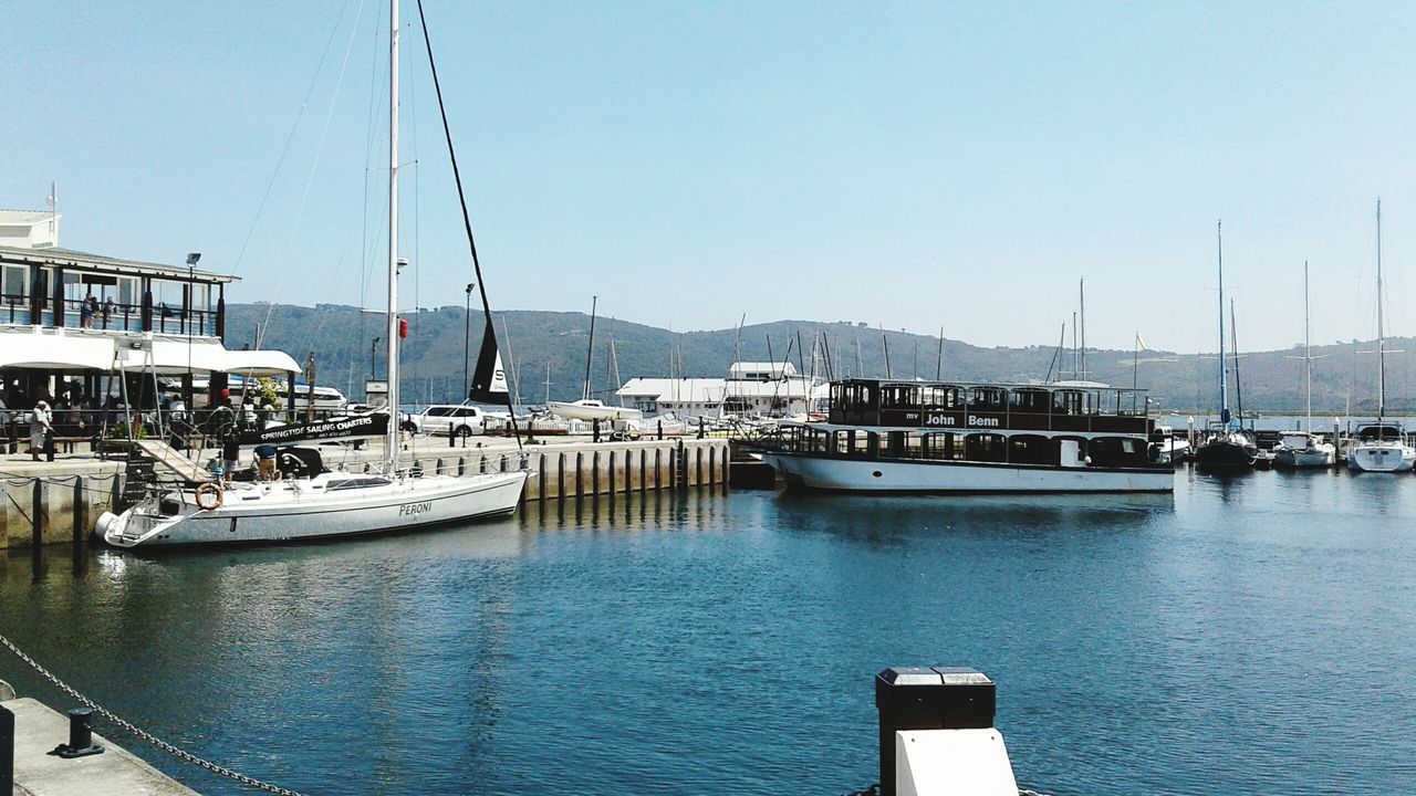 nautical vessel, water, moored, clear sky, boat, transportation, mode of transport, harbor, sea, copy space, mountain, blue, built structure, building exterior, sailboat, mast, waterfront, architecture, day, nature