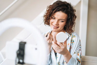 Young woman blogger in home clothes doing makeup with mobile phone and ring lamp online in studio