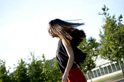 Side view of happy teenage girl tossing hair against sky