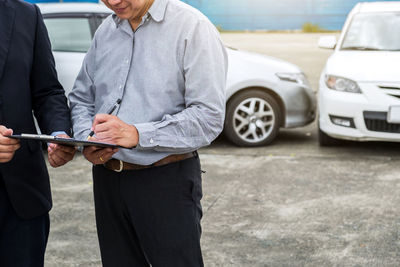 Midsection of man signing paper by insurance agent against cars