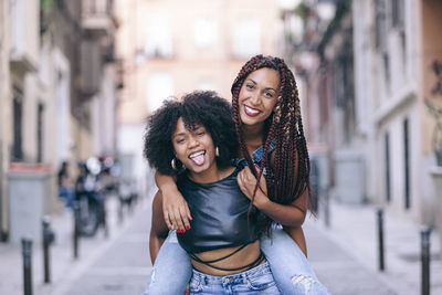 Portrait of playful woman piggybacking friend while standing in city