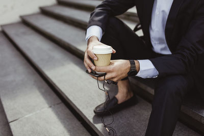 Low section of businessman with disposable cup and smart phone sitting on staircase