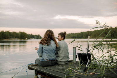 Smiling girl talking to sibling while sitting on pier during sunset