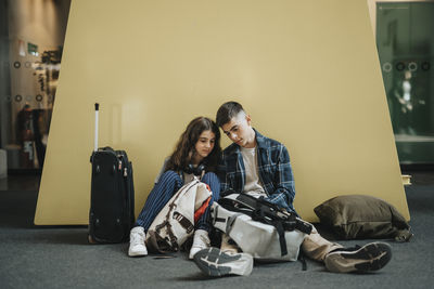 Boy and girl sharing smart phone while sitting with luggage against yellow wall