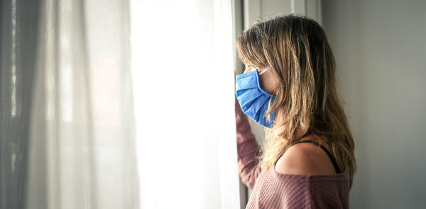 Woman looking outside the window with surgical mask during coronavirus lockdown. covid-19 people