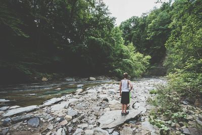 Rear view of man standing on rock at riverbank in forest