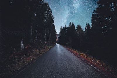 Diminishing perspective of empty road amidst trees in forest against sky at night