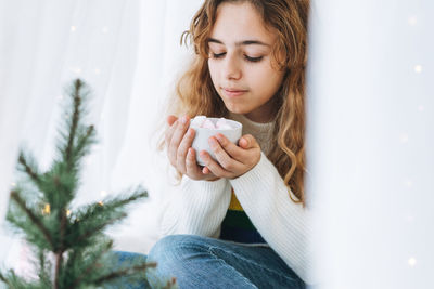 Beautiful teenager girl with cup of cacao with marshmallow sitting on windowsill in cozy room