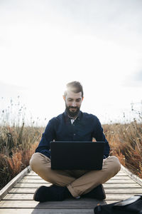 Mid adult man using laptop while sitting on boardwalk against sky