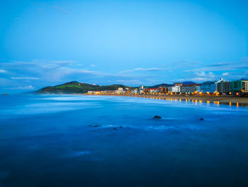 Illuminated buildings by sea against blue sky