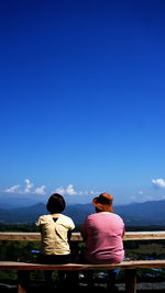 Rear view of friends looking at landscape while sitting on bench against blue sky