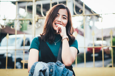 Portrait of smiling young woman biting index finger against fence