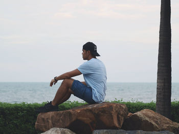 Man sitting on rock looking at sea shore against sky and see sunset