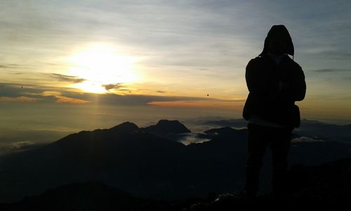 Rear view of person wearing hooded jacket standing on mountain against sky during sunset