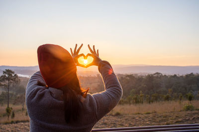 A woman with a heart shaped hand with the sun in the middle of the heart shape.