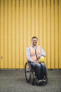 Happy construction worker sitting with hardhat in wheelchair against yellow metal wall