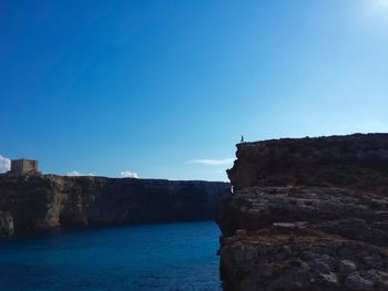 Mid distant view of man on cliff by sea against clear blue sky at comino